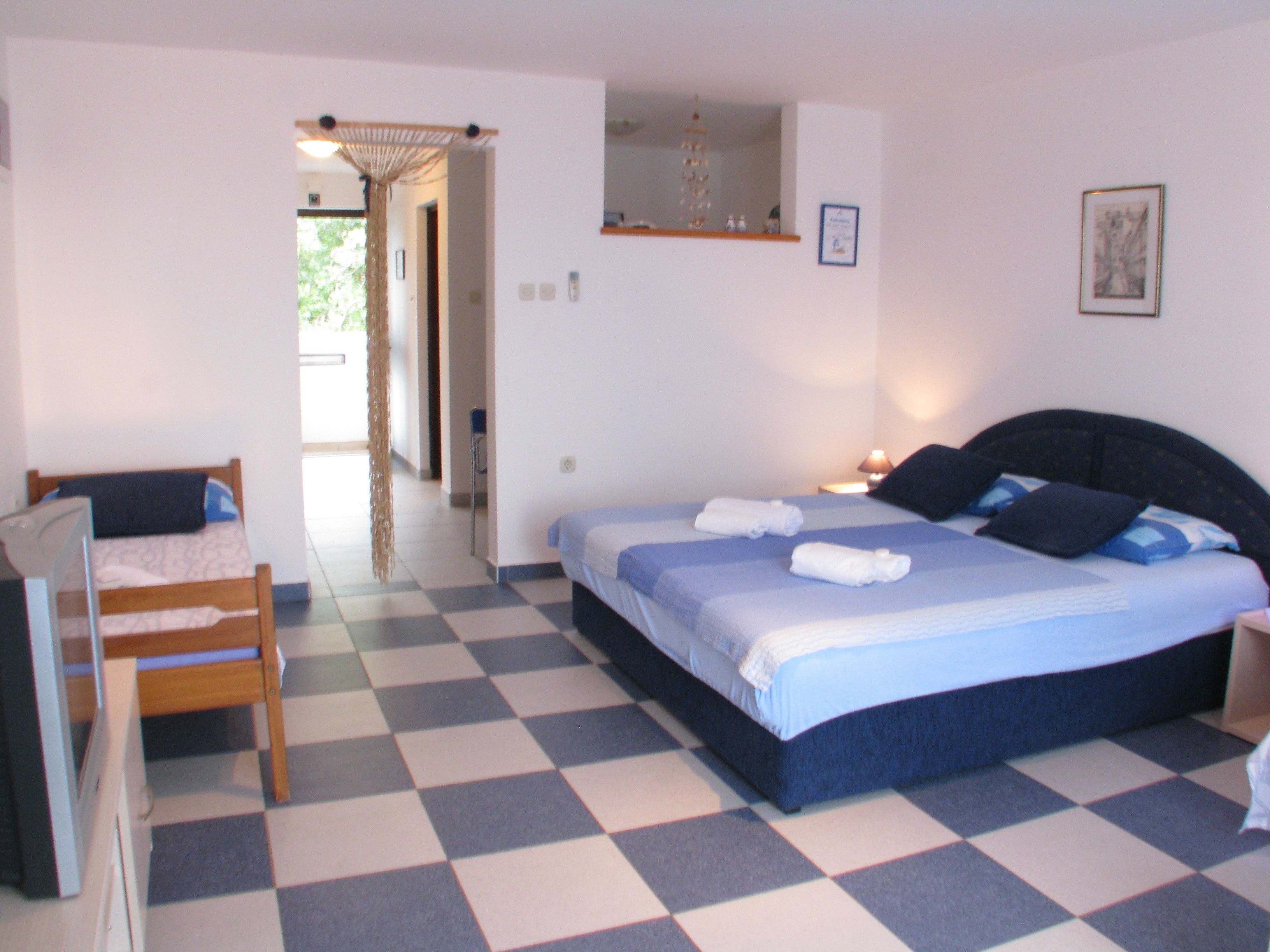 Quarto com piso xadrez, cama com roupa de cama azul, mesa de cabeceira com abajur e uma cadeira.
