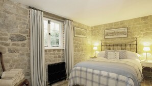 Een slaapkamer met stenen muren, een bed met een geruite plaid, een stoel met handdoeken, een radiator en twee bedlampen.