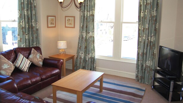 A living room with a brown leather sofa, a wooden coffee table, a TV, and a lamp on a small table. - A spacious apartment with stunning views of Skiddaw and Latrigg. (Keswick)