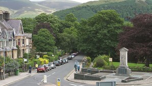 A residential street with parked cars, a statue, and a backdrop of green hills. - A spacious apartment with stunning views of Skiddaw and Latrigg. (Keswick)