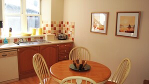 A compact kitchen with wooden cabinets, a sink, and a dining table with four chairs. There are two framed pictures on the wall. - A spacious apartment with stunning views of Skiddaw and Latrigg. (Keswick)