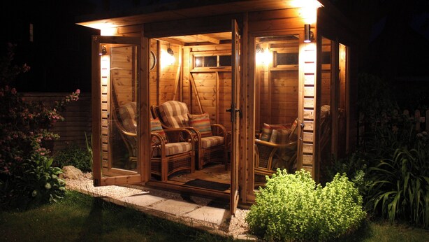 Eine Holzjagdhaus mit Veranda, möbliert mit Korbstühlen und einer Bank, umgeben von einem Garten.