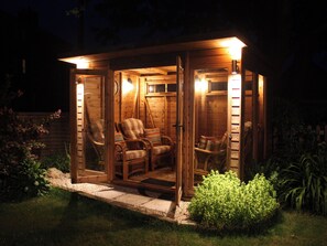 Eine Holzjagdhaus mit Veranda, möbliert mit Korbstühlen und einer Bank, umgeben von einem Garten.