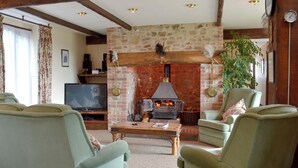 A living room with a stone fireplace, green upholstered chairs, a television, and a wooden ceiling.