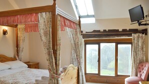 A bedroom with a four-poster bed, a wooden nightstand, a pink chair, a television mounted on the wall, and a large window with a view of greenery.