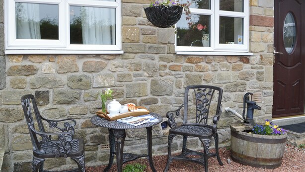 Un bâtiment en pierre avec deux fenêtres, une porte, une table entourée de chaises et un plateau contenant divers objets.