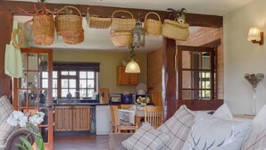 Een gezellige woonkamer met een bank, een salontafel en een vaas orchideeën. Er is een keukenhoek met houten kastjes en een raam.