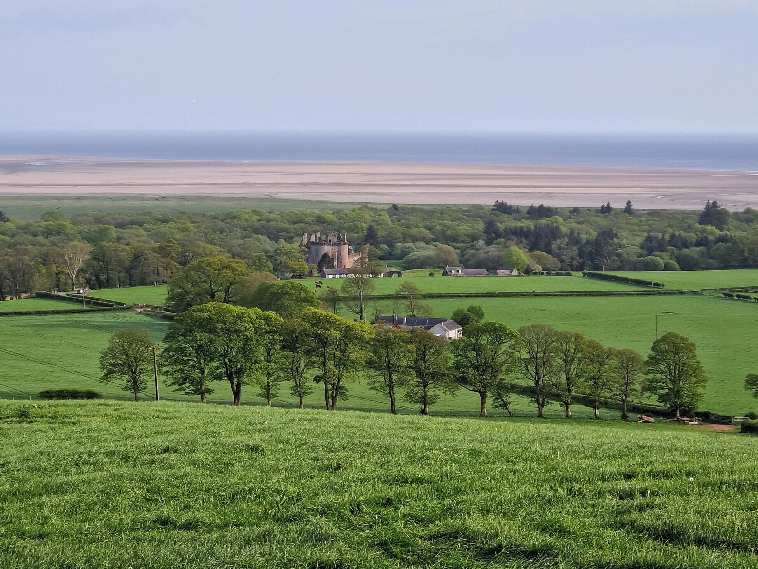 A castle surrounded by green fields and trees, with a view of the sea in the background.