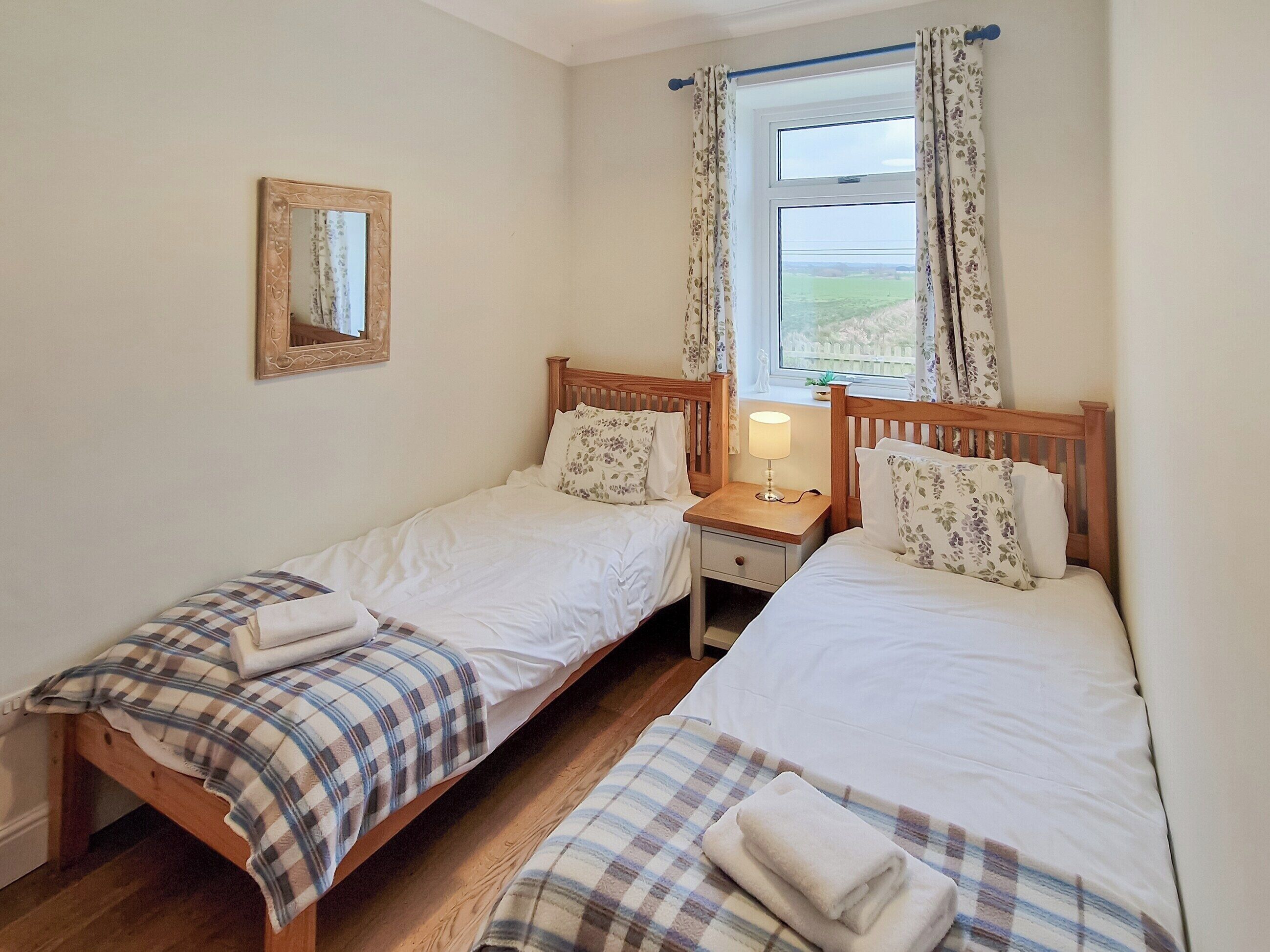 A twin room with two single beds, a bedside table with a lamp, a mirror on the wall, and a window with floral curtains.
