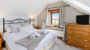 A bedroom with a bed, bedside table, lamp, window with curtains, dresser, and a television.