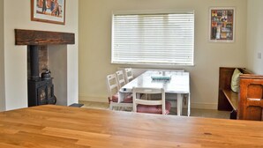 Un coin repas avec une table en bois, des chaises blanches, une cheminée et une fenêtre avec des stores.