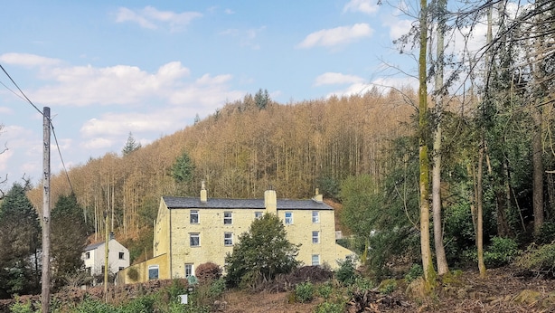 A two-story stone building with multiple windows, surrounded by a forested hillside.
