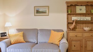 A living room with a grey sofa, two yellow cushions, a wooden cabinet with a sign, and a framed landscape painting on the wall.