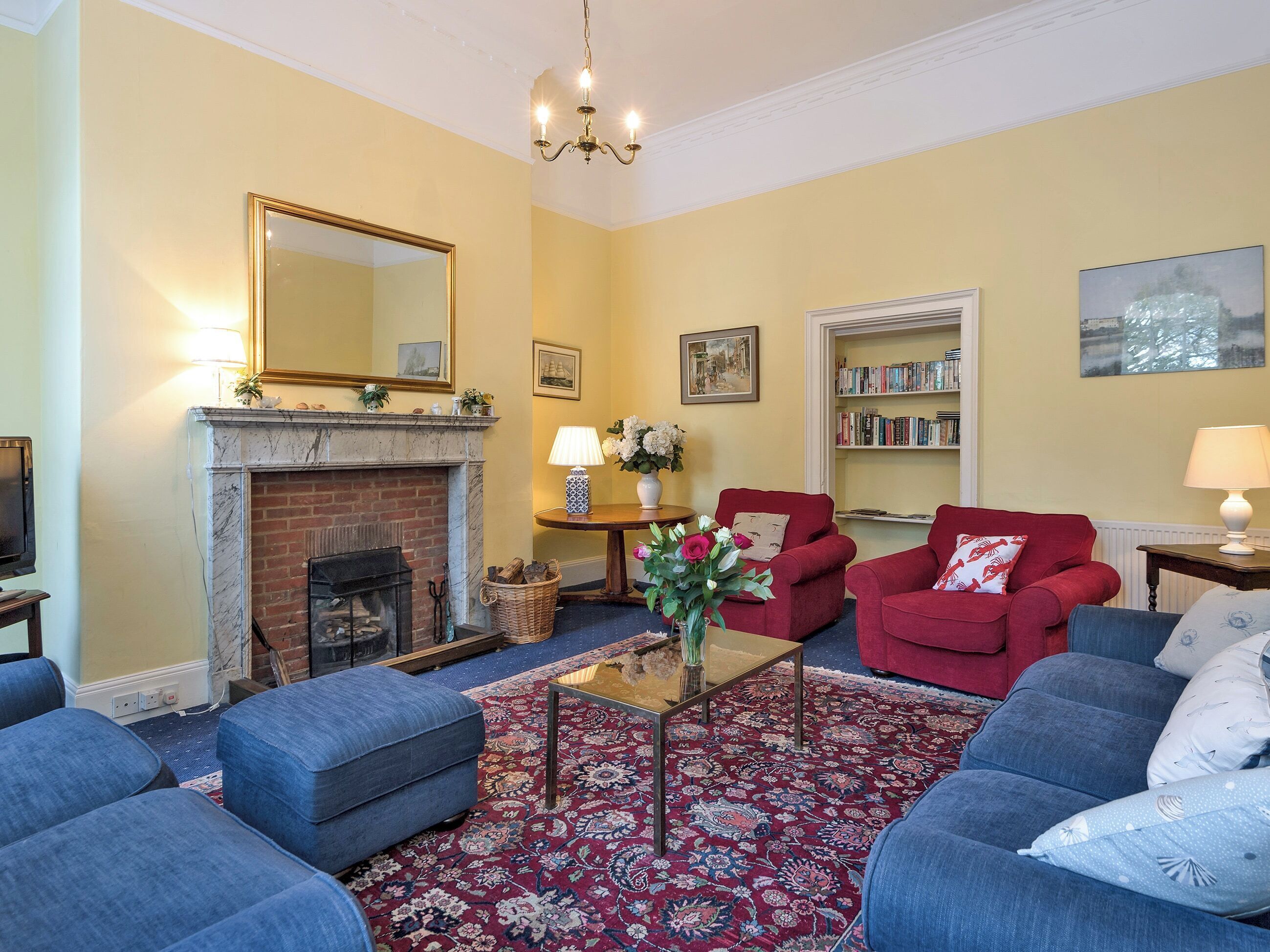 A living room with a fireplace, red armchairs, blue sofas, a coffee table, and a bookshelf.