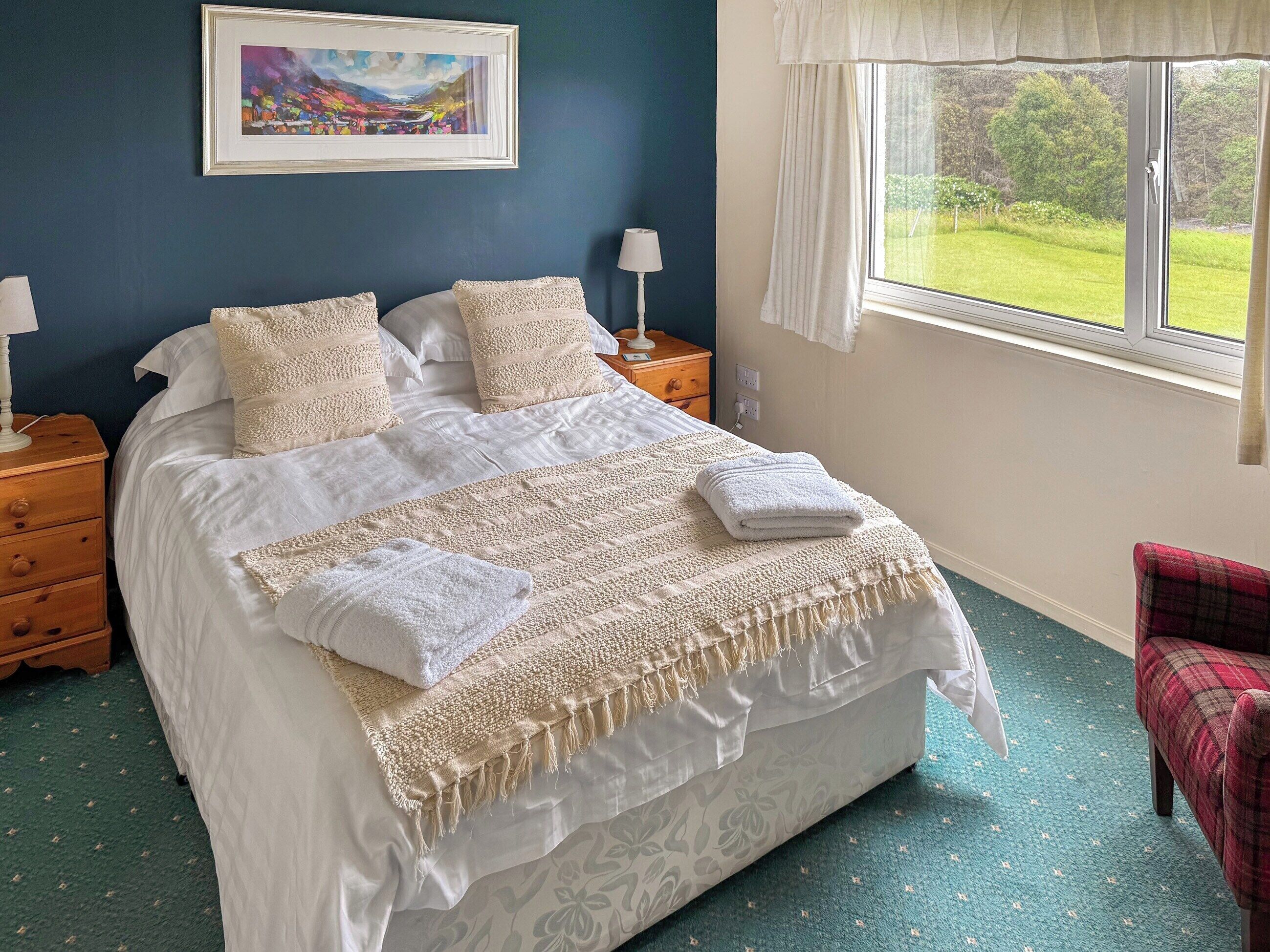 A bedroom with a bed, two bedside tables, a nightstand, a window with a view of greenery, and a framed picture on the wall.