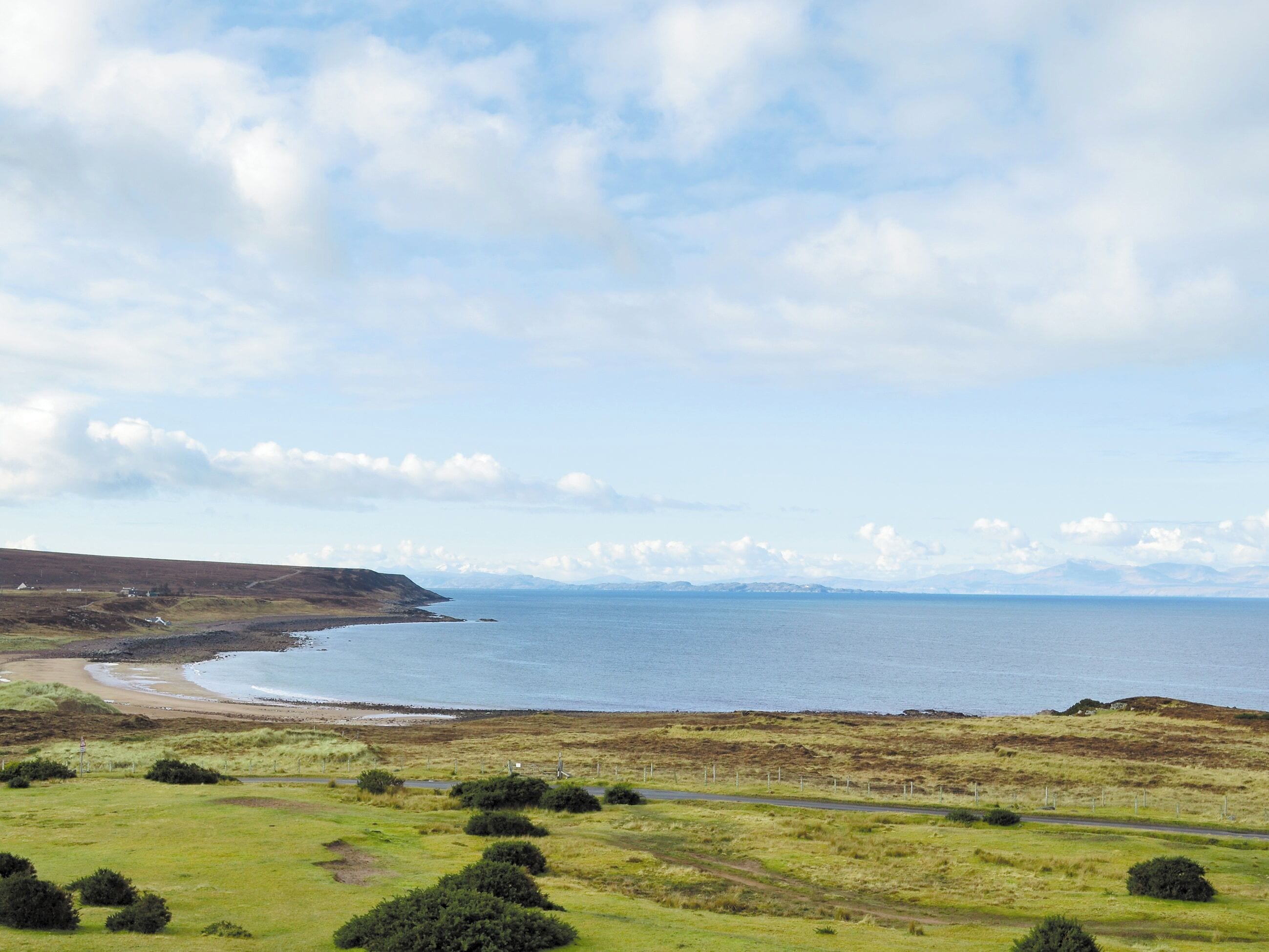 Eine Küstenlandschaft mit einer grasbewachsenen Vordergrundebene, einer Straße und einer entfernten Aussicht auf Meer und Berge.