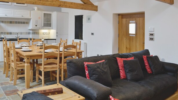 A dining area with a wooden table and chairs, a black sofa with red pillows, and a kitchen in the background. - 2 bedroom accommodation in Tynlon, near Rhosneigr (Tynlon, near Rhosneigr)