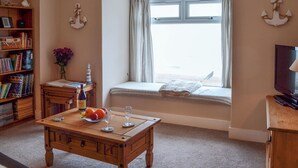 A cozy living room with a wooden coffee table, bookshelves, a TV, and a window with curtains. - 1 bedroom accommodation in Pevensey Bay (Pevensey Bay)