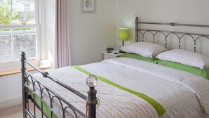 A bedroom with a metal bed frame, a green and white bedspread, a bedside table with a lamp, and a window with curtains. - 2 bedroom accommodation in Buckfastleigh (Buckfastleigh)