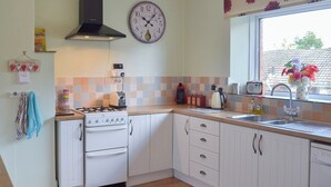 A kitchen with white cabinets, a gas stove, a clock, and a window with a flower vase. - 2 bedroom accommodation in Buckfastleigh (Buckfastleigh)