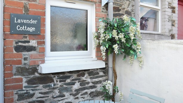 A brick cottage with a sign reading "Lavender Cottage," a window, a white bench, and a potted plant with white flowers. - 2 bedroom accommodation in Buckfastleigh (Buckfastleigh)