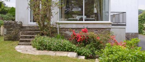 A modern house with a stone staircase leading to a glass door, surrounded by a well-maintained garden with various plants and flowers.