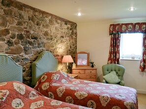 A bedroom with a stone wall, a bed with floral bedding, a dresser, a chair, and a window with floral curtains.