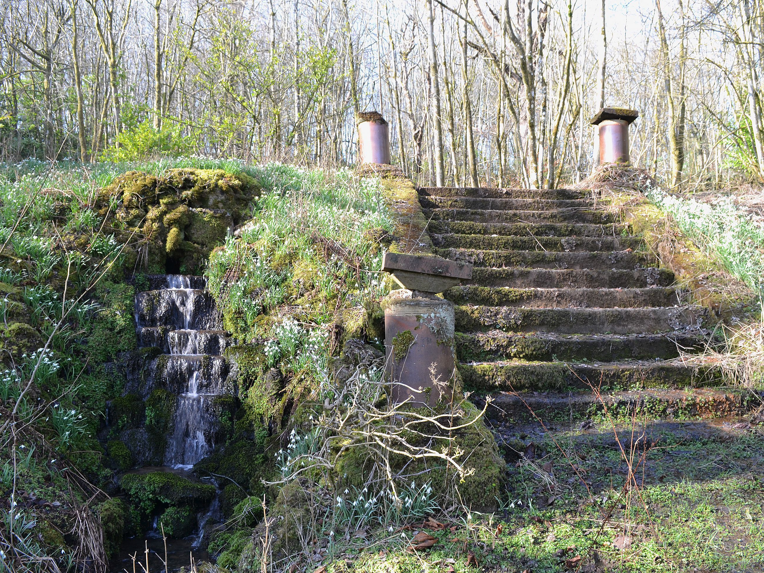 Un escalier en pierre mène à une structure en bois où se trouve une petite cascade.