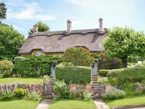 Une maison traditionnelle à toit de chaume, dotée d’un mur en pierre et d’un portail en bois, entourée d’une végétation luxuriante et de plantes en fleurs.