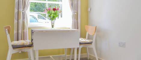 A room with a white table and two chairs, a window with curtains, and a vase with flowers on the table.
