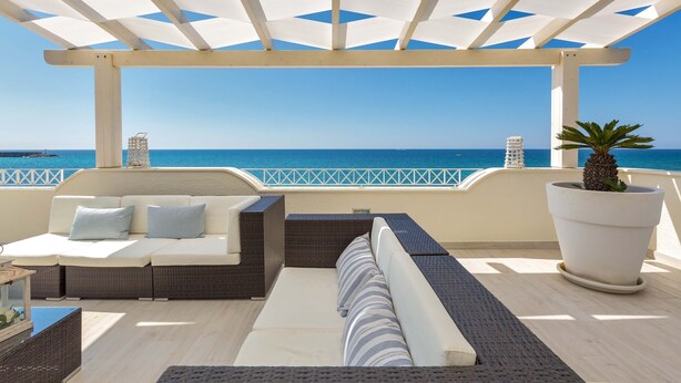 A covered outdoor seating area with a view of the sea, featuring a sofa, cushions, and a large potted plant. - L'Attico di Alessia by Perle di Puglia (Maruggio)