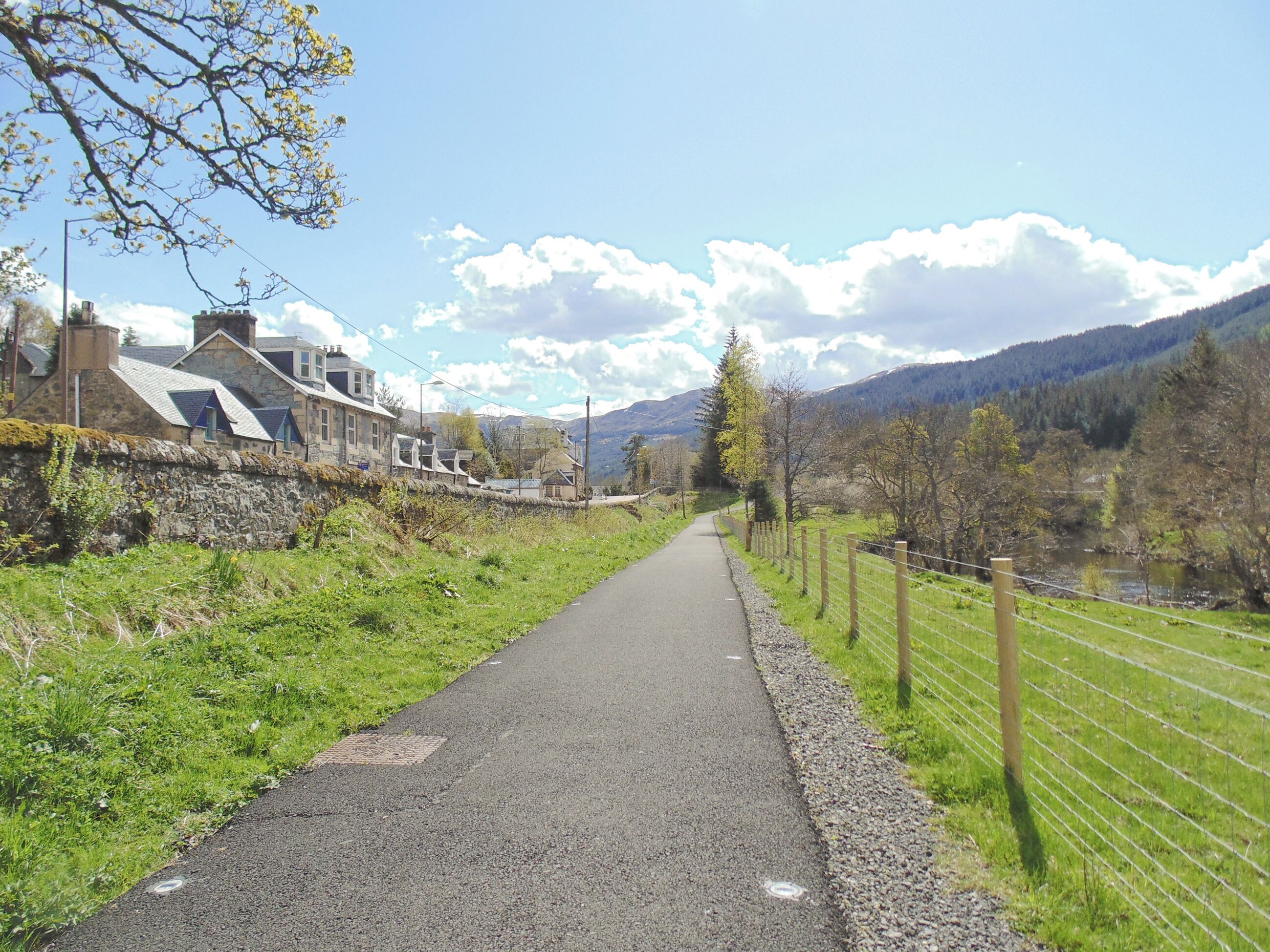 Eine ländliche Straße mit Steinhäusern an einer Seite und einem eingezäunten Feld auf der anderen.