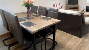 Een eetgedeelte met een houten tafel, zes stoelen, een bloemenvaas, een televisie en een bank.