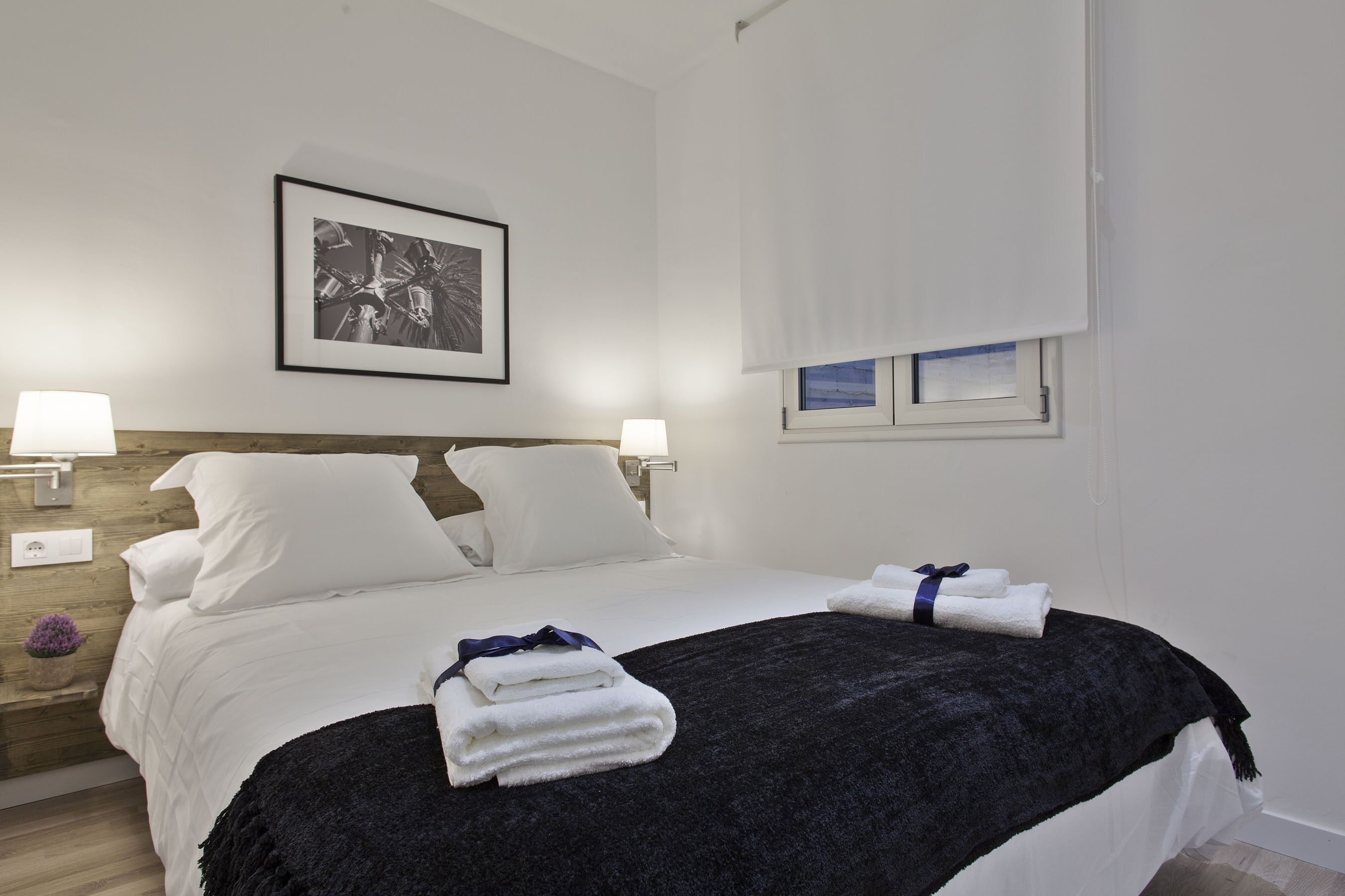 A neatly made bed with white linens, a dark throw blanket, and towels on the side. There is a framed picture on the wall above the bed and a window with a view of the sky.