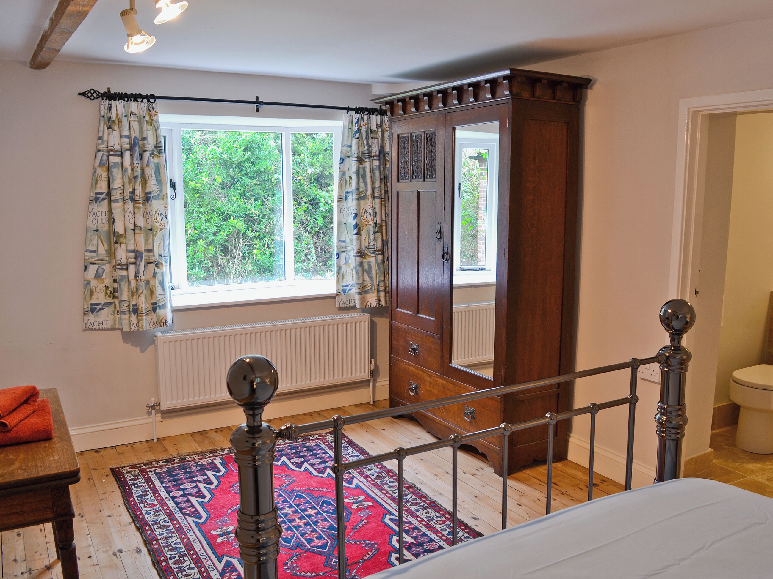 A bedroom with a large window, a wooden wardrobe, a bed, a radiator, and a red patterned rug.