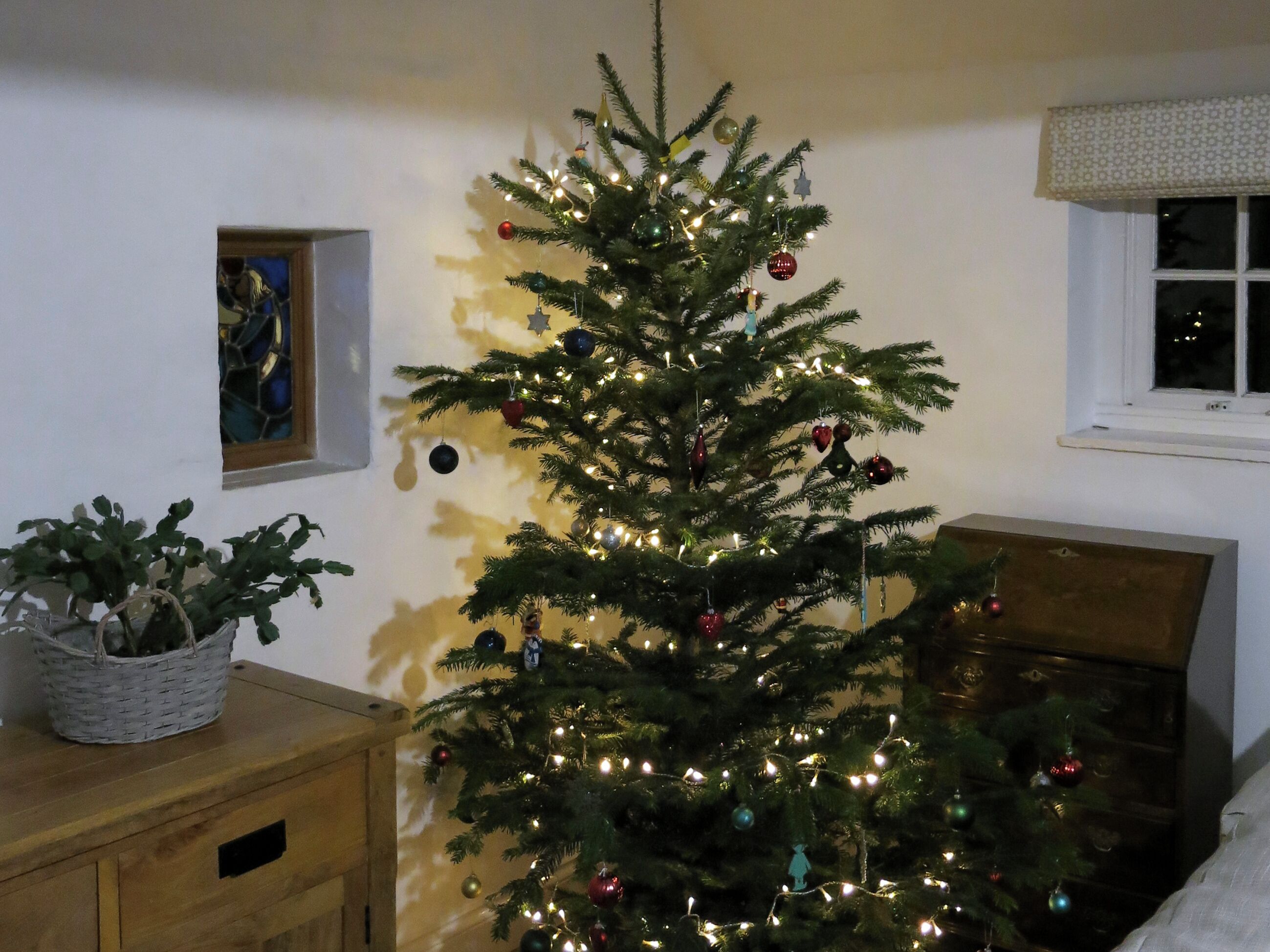A Christmas tree with lights and ornaments in a room with a window and a small painting on the wall.