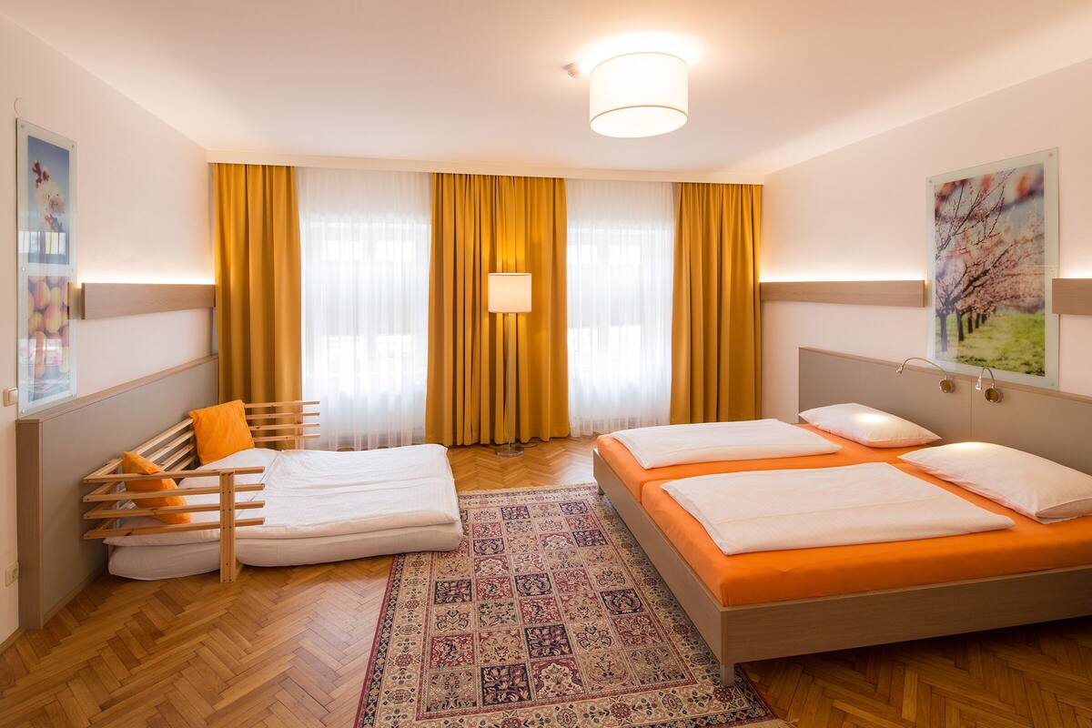 A hotel room with two beds, a wooden bench, a patterned rug, and a large window with curtains.