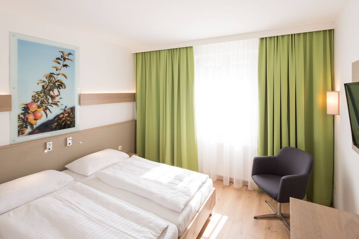 A hotel room with two beds, a desk, a chair, a painting, and green curtains.