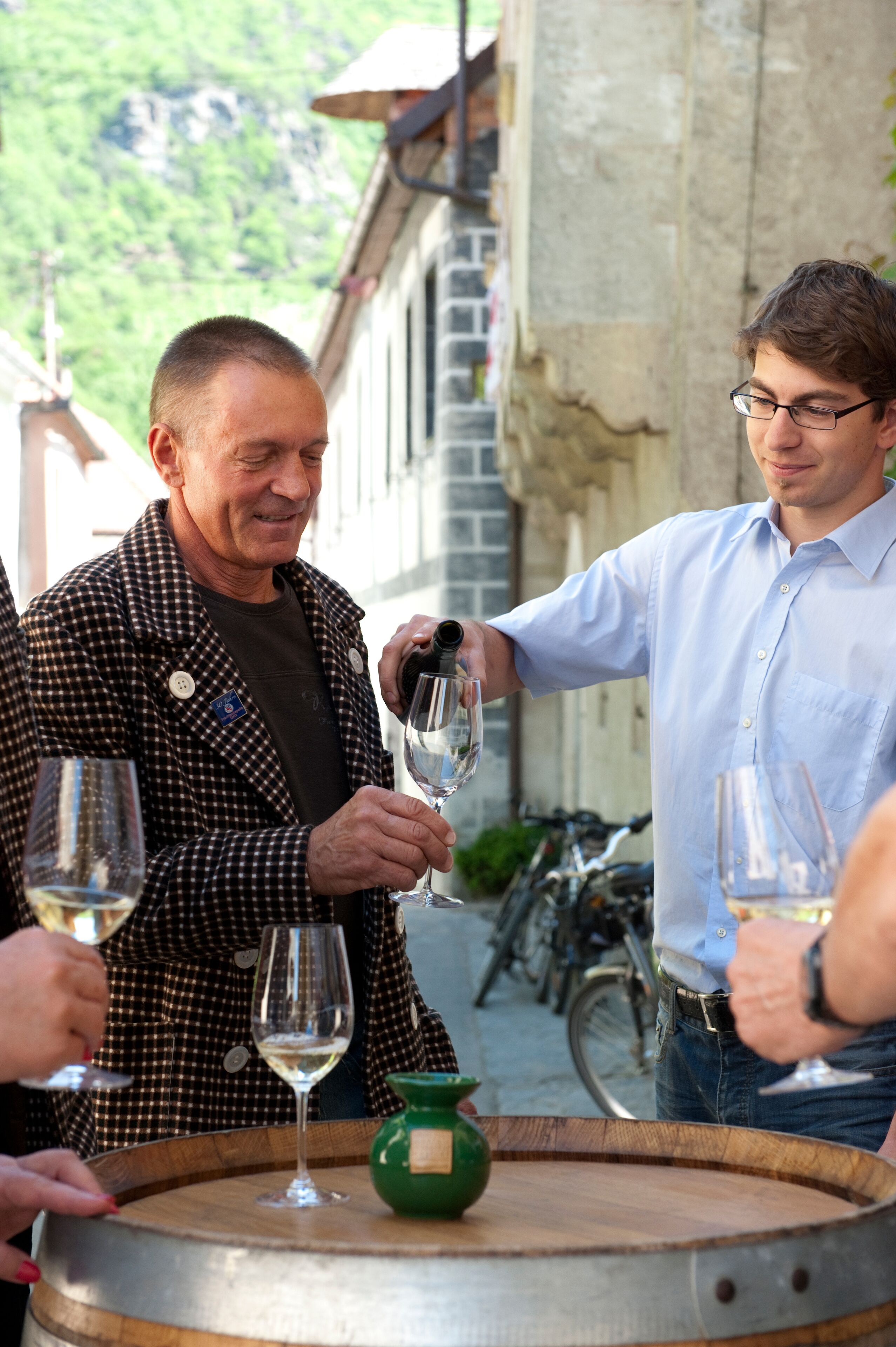 Two people pouring wine from a barrel into glasses, with bicycles in the background.