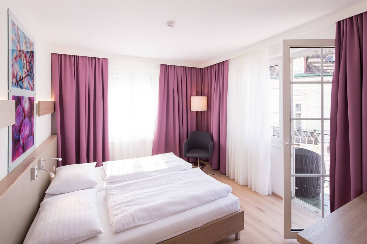 A hotel room with a large bed, a chair, and a balcony with a view.