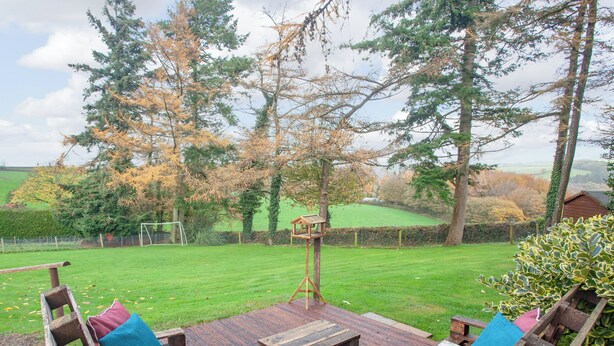 A wooden deck with two benches and a table, overlooking a grassy yard with trees and a distant view of a field. - Challette At Timbertops (Washfield, near Tiverton)
