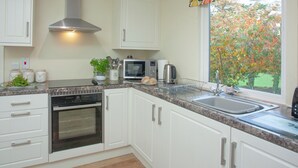 A modern kitchen with white cabinets, stainless steel appliances, and a window with a colorful fruit pattern valance. - Challette At Timbertops (Washfield, near Tiverton)