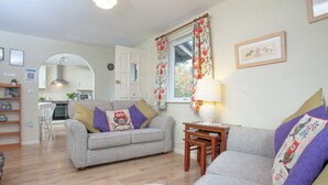 A living room with a grey sofa, a wooden side table with a lamp, and a window with floral curtains. - Challette At Timbertops (Washfield, near Tiverton)