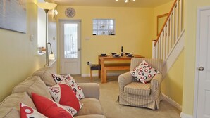 Un salon comprenant un canapé, un fauteuil et une table à manger avec des chaises. On y trouve un escalier avec une rampe en bois et une porte blanche.