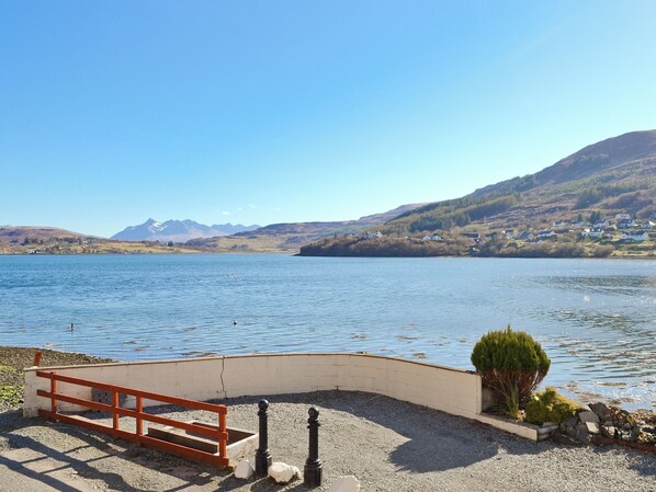A lakeside view with a bench, a small garden, and a distant mountain range.