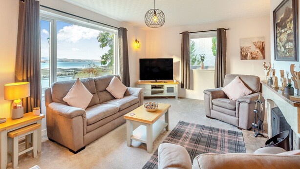 Ett vardagsrum med en soffa, fåtölj och TV, stora fönster med gardiner, en öppen spis och utsikt över havet.