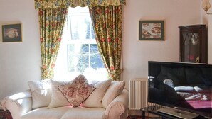A living room with a white sofa, a flat-screen TV, floral curtains, and a radiator. - 5 bedroom accommodation in Byrness Village, near Otterburn (Byrness Village, near Otterburn)