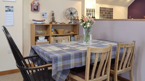 Un coin repas avec une table dressée pour quatre personnes, entourée de chaises en bois. On y trouve un ventilateur, un vase avec des fleurs et une étagère contenant divers objets, notamment une horloge et des objets décoratifs.