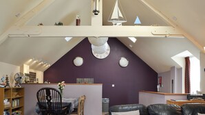 Un espace de vie de style loft avec un plafond haut, des poutres apparentes, un coin repas avec une table et des chaises, un canapé en cuir noir et une maquette de petit voilier.