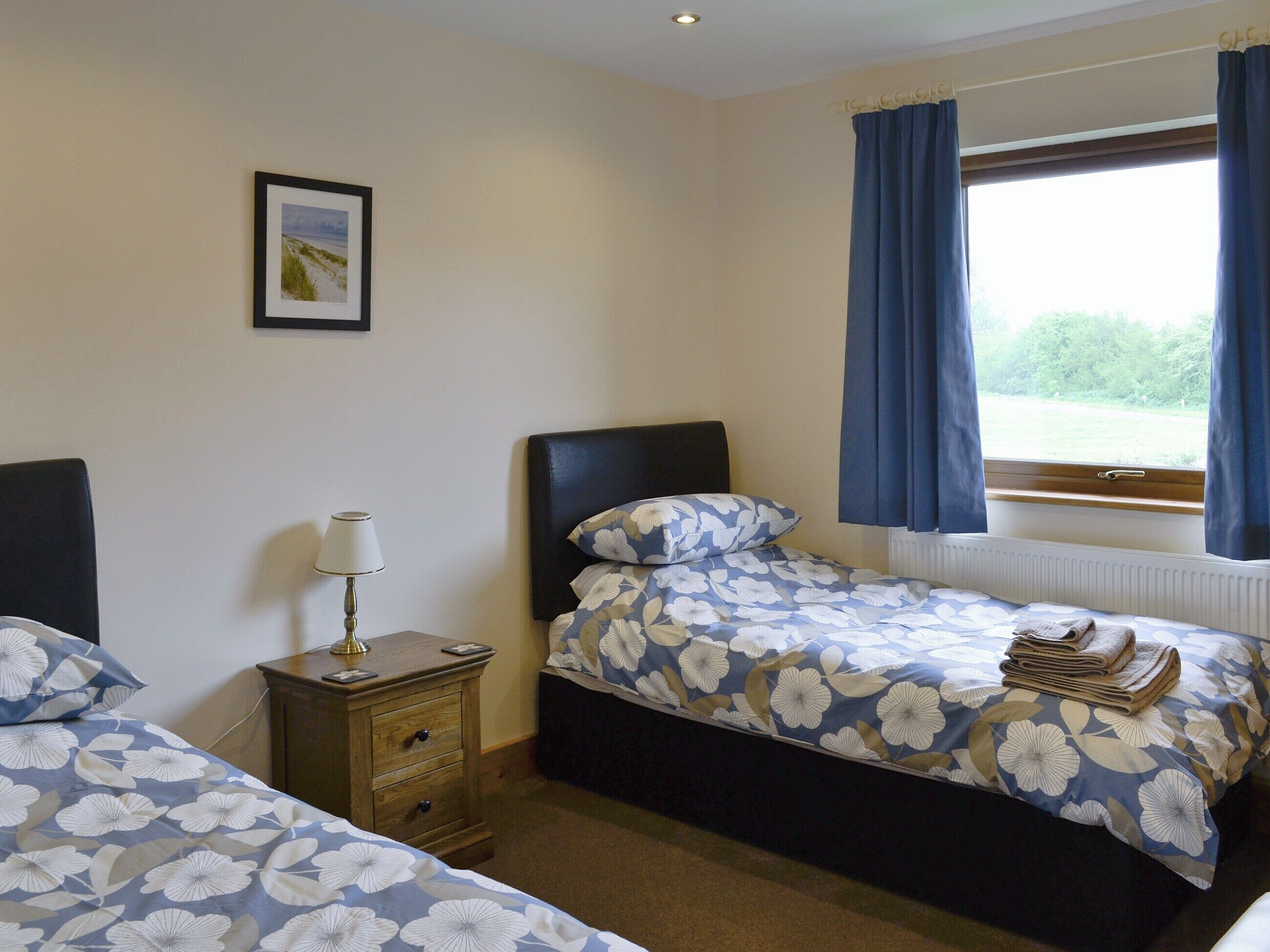 A bedroom with two single beds, a nightstand with a lamp, a framed picture on the wall, and a window with curtains.